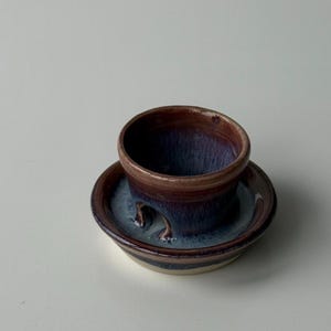 May include: A handcrafted ceramic cup and saucer set. The cup is a deep brown with a purple interior, and the saucer has a matching colour scheme. The set is glazed with a glossy finish, and the design is simple and elegant.