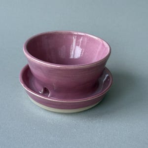 May include: A small, round, ceramic bowl with a glossy, deep mauve glaze on the interior and exterior, resting on a matching saucer. The saucer features a small, circular cutout on its side, and the base of the bowl and saucer is unglazed.