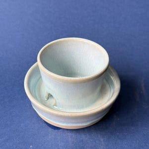 May include: A light blue ceramic cup and saucer set with a glossy finish and subtle tan rim detailing. The cup features a small arched opening at the base, and the saucer has a matching light blue glaze with a smooth, rounded edge design.