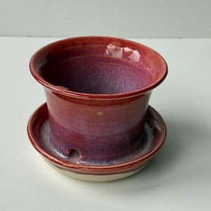 May include: A handcrafted ceramic plant pot in red and purple hues, with a matching saucer. The pot features a flared rim and a drainage hole. The saucer is round with a raised edge.