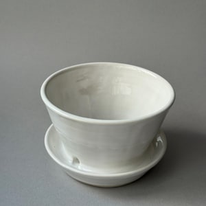 May include: A white ceramic planter bowl with a matching saucer base. The bowl features a wide, flared rim and a smooth, glossy finish. A small drainage hole is visible near the base where the bowl connects to the integrated saucer component.