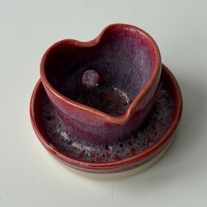 May include: A heart-shaped ceramic soap dish with a round base, glazed in red and purple hues. The soap holder has a small drainage hole. The base is white. A handcrafted item for the bathroom.