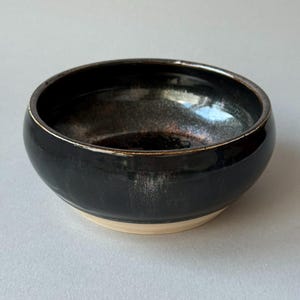 May include: A small, round ceramic bowl with a glossy black glaze on the interior and exterior. The rim features a subtle metallic sheen, while the base remains unglazed, revealing a light beige, matte clay texture at the bottom edge.