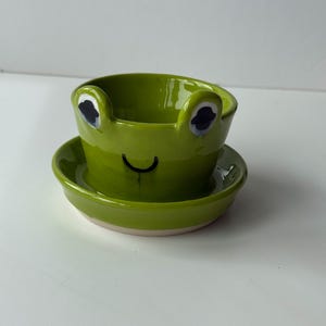 May include: A green ceramic frog-shaped planter with a matching saucer. The planter features a smiling face with black eyes and a curved mouth. Ideal for small plants or succulents, this whimsical item adds a touch of fun to any space. The planter is a decorative item.