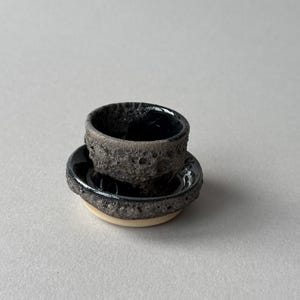 May include: A small ceramic cup and matching saucer set featuring a dark, porous, and textured exterior surface. The interior of the cup and the centre of the saucer have a smooth, glossy black finish, while the base of the saucer is light beige.