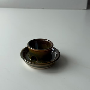 May include: A small, handcrafted ceramic candle holder with a dark brown interior and a green and brown exterior. The candle holder has a circular base with a small, round cup in the centre to hold a candle. The design is simple and elegant.