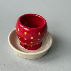 May include: A small, red, rounded ceramic vessel with yellow polka dots, featuring a hollow interior and a small opening at the base, resting inside a circular, off-white ceramic saucer with a smooth, glazed finish and a raised rim.