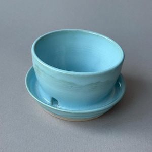 May include: A light blue ceramic planter with a matching saucer. The bowl-shaped planter features a smooth, glossy finish with subtle variations in colour and a small drainage hole visible at the base where it rests on the circular saucer.