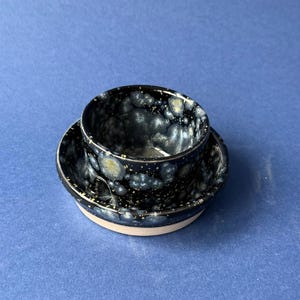 May include: A small ceramic cup and saucer set featuring a dark navy blue glaze with a speckled, crystalline pattern in shades of white, grey, and pale yellow. The base of the saucer shows a smooth, unglazed light beige ceramic rim.