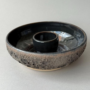 May include: A circular ceramic bowl with a central cylindrical cup. The interior features a smooth, glossy black glaze, while the exterior displays a textured, cratered surface in dark grey and brown tones with a light beige base rim.