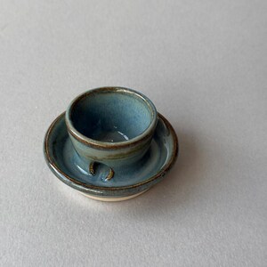 May include: A small, round, blue-grey ceramic cup and saucer set. The cup features a smooth, glossy glaze with darker brown-toned edges and a small, arched cutout at the base. The saucer has a matching blue-grey finish with a visible light-coloured rim.