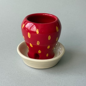 May include: A red ceramic strawberry-shaped planter with yellow seed details and a drainage hole at the base, resting on a circular off-white saucer. The glossy finish reflects light, highlighting the rounded form of the small plant pot.