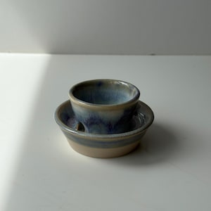 May include: A small, handmade ceramic soap dish with a matching cup. The dish is a light beige colour with a blue and purple glaze around the rim. The cup is blue and purple. The set is designed to hold a bar of soap.