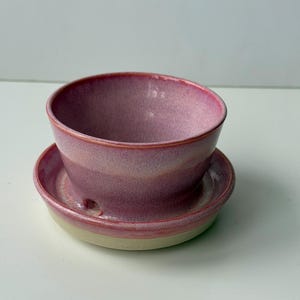 May include: A handmade ceramic bowl and saucer set. The bowl and saucer are a gradient of pink hues, with a lighter base. The bowl has a rounded shape and sits atop a matching saucer. The set is ideal for serving small portions.