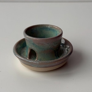 May include: A handmade ceramic match holder with a teal and brown glaze. The holder has a small cup shape with a hole on the side, set on a matching saucer base. The pottery has a rustic, artisanal appearance.
