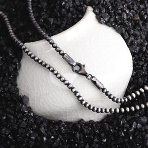 Pode incluir: Um colar de contas de prata com fecho lagosta, marcado "925". O colar é exibido em um objeto branco de formato orgânico, em um fundo de pedras pretas. As contas são pequenas e espaçadas uniformemente.