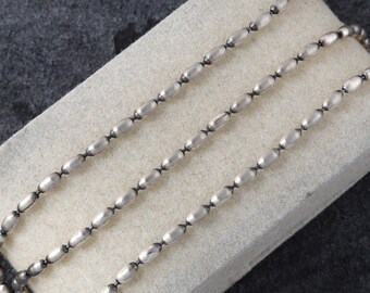 May include: A silver-colored beaded necklace with oval-shaped beads connected by small links. The necklace is displayed on a light-colored, rectangular surface against a dark gray background. The beads have a polished finish.