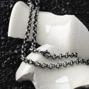 Puede incluir: Un collar de cadena plateada con cierre de mosquetón. El collar presenta una serie de pequeños eslabones circulares. La cadena se muestra sobre una superficie blanca texturizada sobre un fondo oscuro.