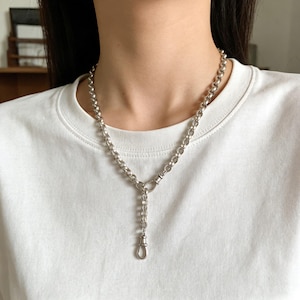 Puede incluir: Collar plateado con un diseño de cadena gruesa. El collar presenta una caída en forma de Y con una cadena más pequeña y un cierre de broche. La joya se usa sobre una camiseta blanca.