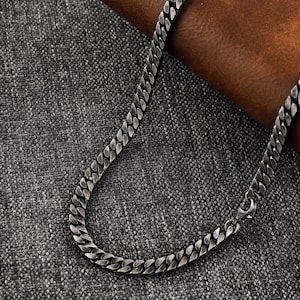 Op de afbeelding: Een zilveren gourmetketting met een karabijnsluiting. De ketting heeft een plat, in elkaar grijpend schakelontwerp. De ketting wordt gepresenteerd op een grijze, getextureerde ondergrond met een bruine lederen achtergrond.