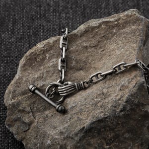 Puede incluir: Un collar plateado con un cierre de palanca único. El cierre presenta un diseño de mano que sostiene un anillo, conectado a una cadena con un cierre de barra. El collar se muestra sobre una superficie rocosa texturizada.
