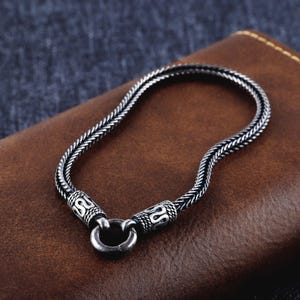 Puede incluir: Una pulsera plateada con un diseño de cadena trenzada. La pulsera presenta cierres decorativos con un patrón intrincado y un cierre circular. La joya se exhibe sobre una superficie de cuero marrón.
