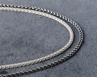 May include: Close-up of two silver-toned chain necklaces. One is a bright silver color, and the other is a darker, oxidized silver. The chains have a woven, wheat-style design. The background is a muted gray.