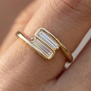 May include: A gold ring with a unique design. The ring features two rectangular, clear stones set in a curved, open-style band. The band is a bright gold color, and the stones are set side-by-side, creating a modern and elegant look. The ring is shown on a finger.