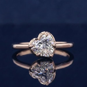 May include: A rose gold ring with a heart-shaped diamond. The diamond is clear and faceted, reflecting light. The ring is set against a dark blue background, with a reflection of the ring visible below.