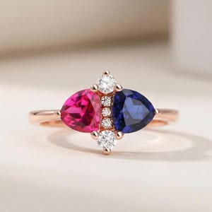 May include: A rose gold ring with a striking design. It features a fuchsia pink and a deep blue gemstone, accented by small, sparkling white stones. The ring's band has a delicate, polished finish.