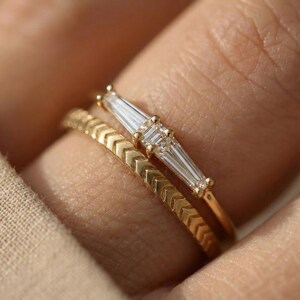 May include: Two gold rings. The top ring features a baguette-cut diamond design. The bottom ring has a chevron pattern. The rings are set against a blurred background.