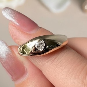 Puede incluir: Un anillo dorado con un diamante en forma de corazón. El anillo tiene un diseño liso y redondeado. El anillo lo sostiene una persona con esmalte de uñas rosa y blanco. Una flor blanca y un objeto dorado están en el fondo.