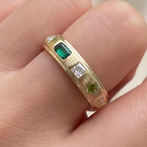 May include: A gold ring with an emerald-cut green gemstone, a square white gemstone, and a round green gemstone. The ring is set with small diamonds and has a brushed finish.
