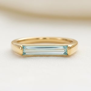 May include: A gold ring with a rectangular, light blue gemstone. The ring has a simple, elegant design with a polished finish. The gemstone is set horizontally in the center of the ring.