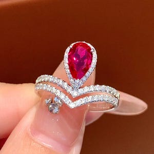 Op de afbeelding: Een zilveren ring met een peervormige, levendige rode edelsteen in het midden, omringd door een halo van kleinere, heldere stenen. De ringband heeft een dubbellaags, chevron-achtig ontwerp, eveneens versierd met kleine, heldere stenen. De ring wordt door een persoon vastgehouden.