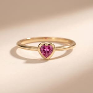 Può includere: Un anello d'oro con una gemma magenta a forma di cuore. La fascia dell'anello è sottile e lucida, e la gemma è incastonata in una lunetta. L'anello è presentato su uno sfondo neutro.