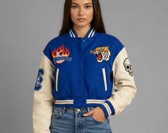 May include: Blue and cream varsity jacket with embroidered patches. Features a tiger, skull, and flame design. The jacket has a snap-button closure and striped cuffs.