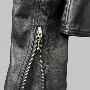 May include: Close-up of a black leather jacket sleeve with a silver zipper. The zipper runs along the sleeve's edge, and the leather has a textured appearance. The jacket appears to be new.