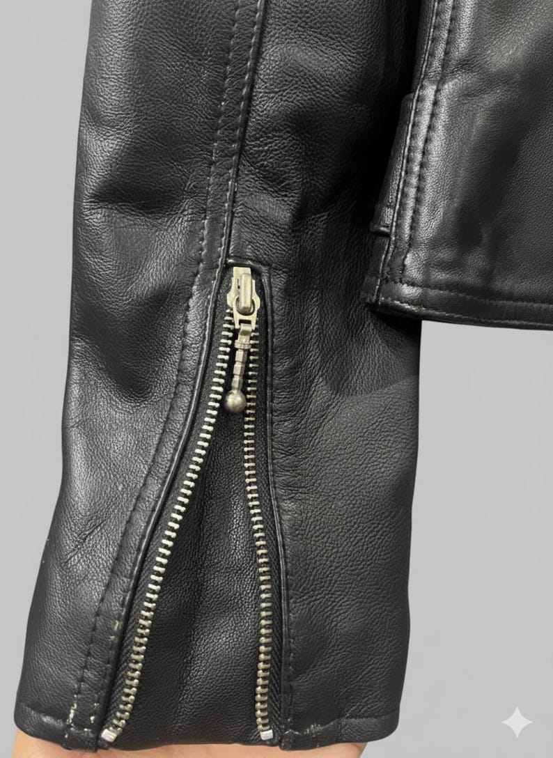 May include: Close-up of a black leather jacket sleeve with a silver zipper. The zipper runs along the sleeve's edge, and the leather has a textured appearance. The jacket appears to be new.