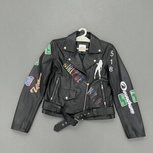May include: Black faux leather jacket with a belted waist and multiple colorful patches. The jacket features the text "GIRLZ BE WYLD" and a silhouette of a woman. The jacket is hanging on a white hanger.