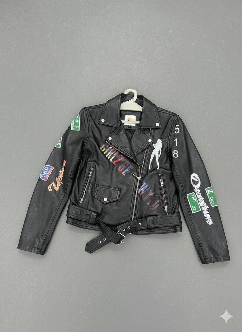 May include: Black faux leather jacket with a belted waist and multiple colorful patches. The jacket features the text "GIRLZ BE WYLD" and a silhouette of a woman. The jacket is hanging on a white hanger.