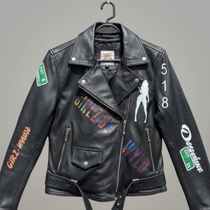 May include: Black leather jacket with a classic biker style, featuring a notched collar, asymmetrical zip closure, and a belted waist. The jacket is adorned with colorful graffiti-style text and a white silhouette graphic. Includes Exit 6, 23 and 9 signs.