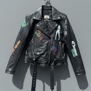 May include: Black leather jacket with a classic biker style, featuring a notched collar, asymmetrical zipper, and a belted waist. The jacket is adorned with colorful graffiti-style graphics and the number 518.