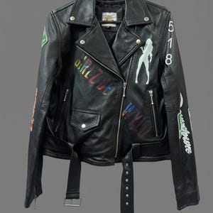 May include: Black leather jacket with a classic biker style, featuring a notched collar, asymmetrical zip closure, and a belted waist. The jacket is adorned with colorful graphics and text, including the words "GIRL BE" and "WYLD".