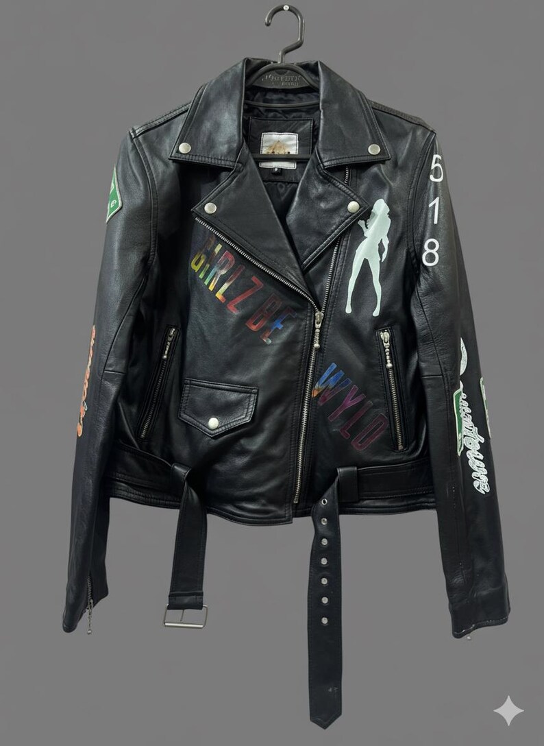 May include: Black leather jacket with a classic biker style, featuring a notched collar, asymmetrical zip closure, and a belted waist. The jacket is adorned with colorful graphics and text, including the words "GIRL BE" and "WYLD".