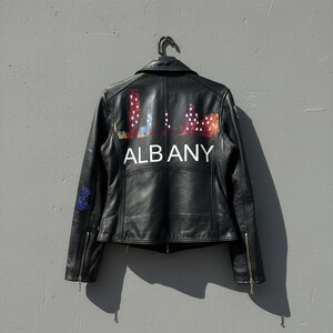 May include: Black leather jacket with a colorful Albany skyline design on the back. The jacket has a classic biker style with a zipper closure and a small graphic on the sleeve. The word "ALBANY" is printed in white.