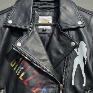 May include: Black leather jacket with a silver zipper and snap closures. The jacket features the text "GIRLZ BE" in rainbow colors and a white silhouette of a woman. The jacket is hanging on a black hanger.