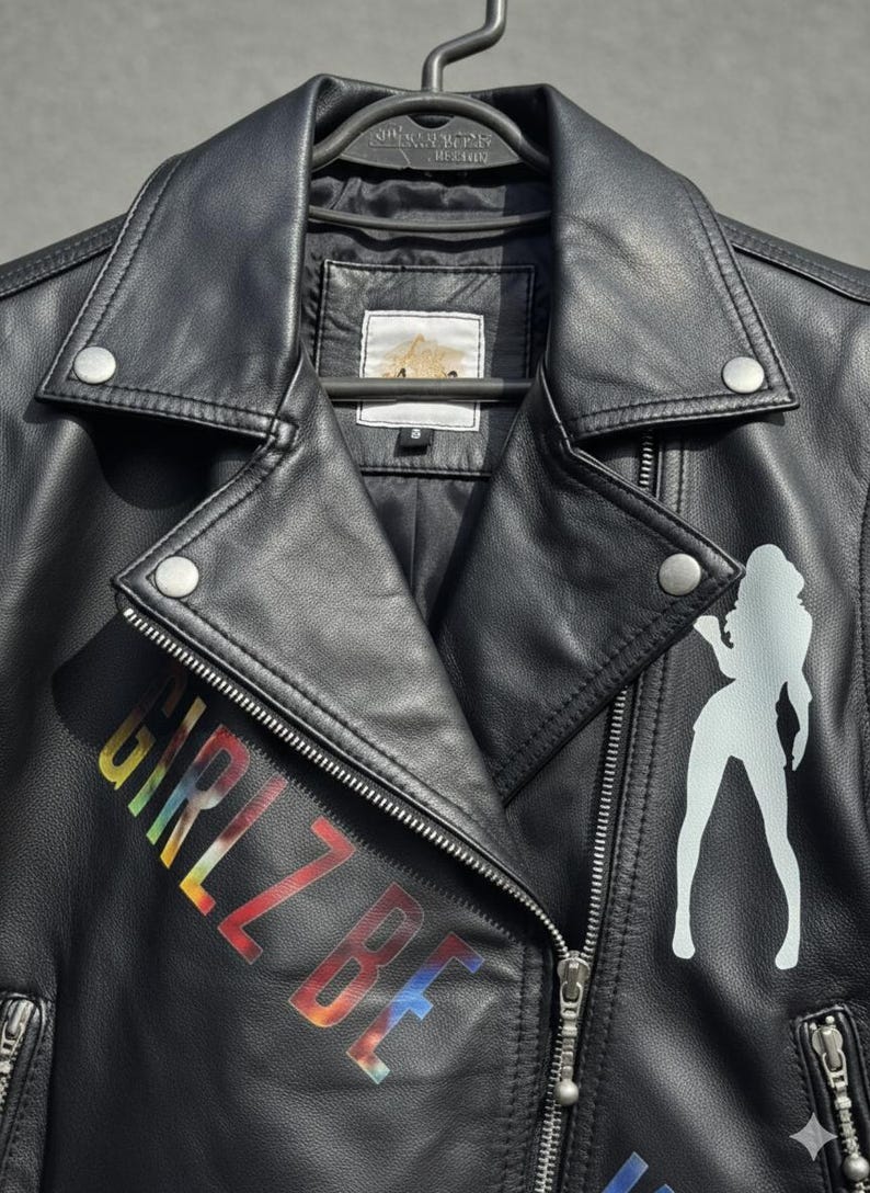 May include: Black leather jacket with a silver zipper and snap closures. The jacket features the text "GIRLZ BE" in rainbow colors and a white silhouette of a woman. The jacket is hanging on a black hanger.