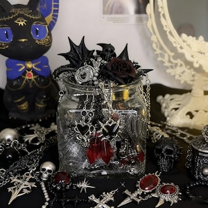 Op de afbeelding: Een doorzichtige glazen pot gevuld met diverse sieraden, waaronder zilverkleurige kettingen, zwarte hartvormige hangers, rode kristallen druppels en metalen kruisbedels. De pot is versierd met zwarte rozenaccenten en kleine schedelfiguren.