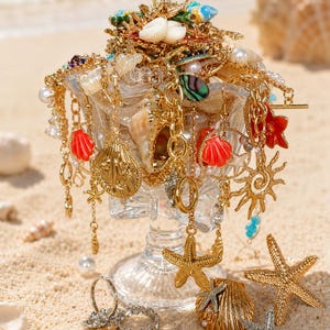 Puede incluir: Un cuenco de cristal transparente sobre pedestal lleno de pulseras y collares con dijes dorados que presentan motivos de conchas, estrellas de mar y soles. La joyería incluye detalles en esmalte rojo y verde, perlas blancas y texturas metálicas.